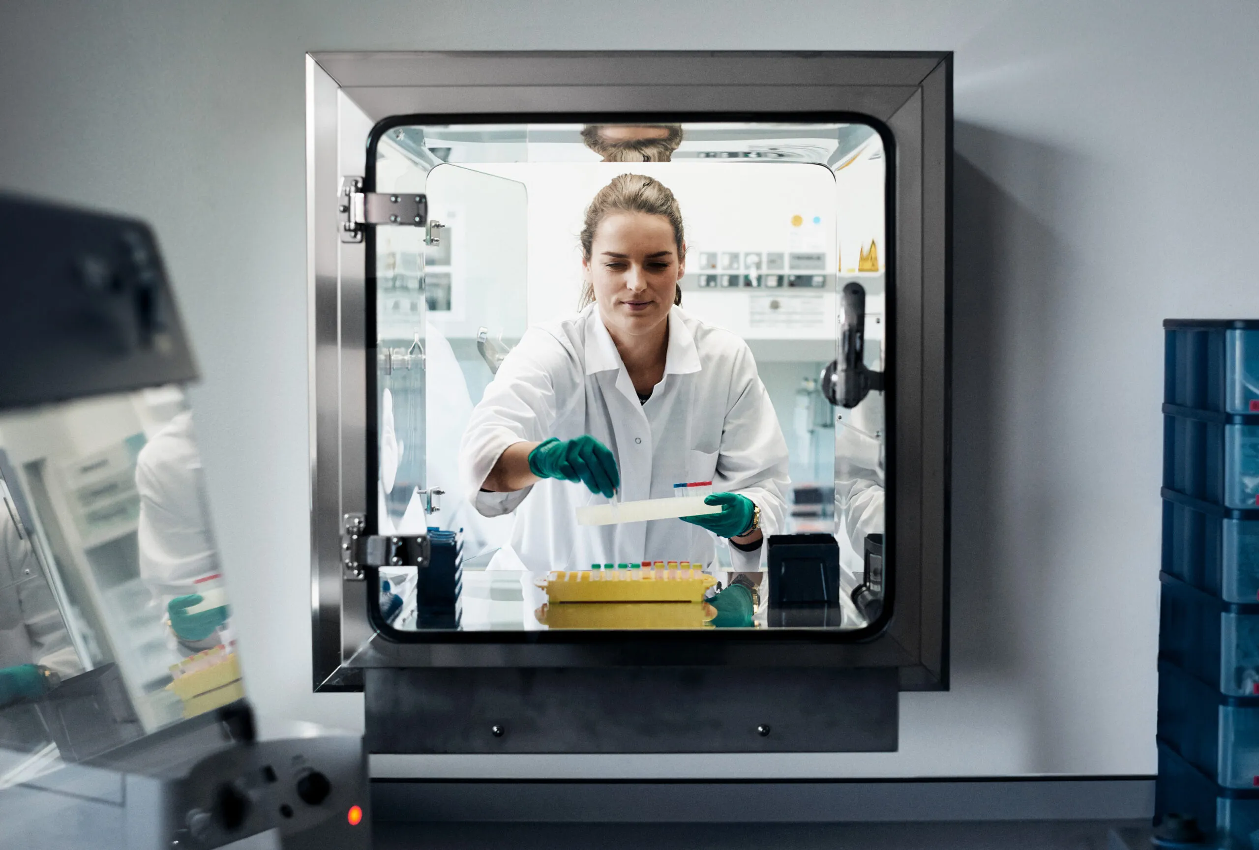 A female laboratory worker in a white coat and green gloves is seen through a window placing laboratory samples into a tray